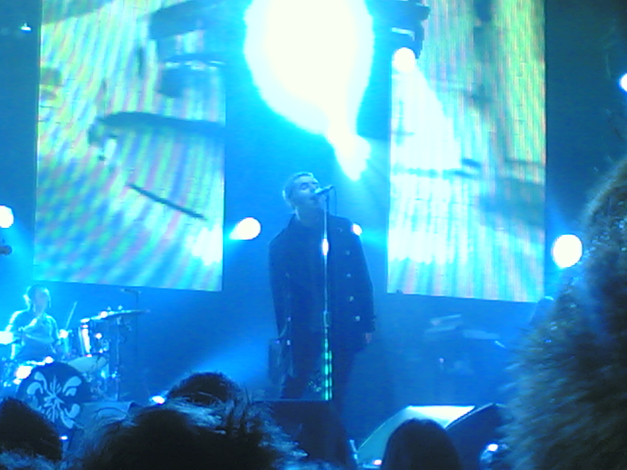 Liam Gallagher singing into a microphone with his hands behind his back on stage in 2009